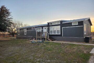 A gray modular home with white trim sits in a grassy yard at sunset. Two bikes are on the ground in front, and trees are visible in the background.