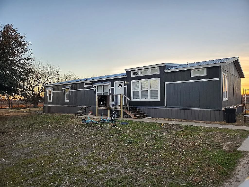 A gray modular home with white trim sits in a grassy yard at sunset. Two bikes are on the ground in front, and trees are visible in the background.
