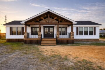 A single-story house with a modern rustic design features a central porch with wooden beams and stone accents. The white exterior and dark shutters create a cozy, welcoming look under a partly cloudy sky.
