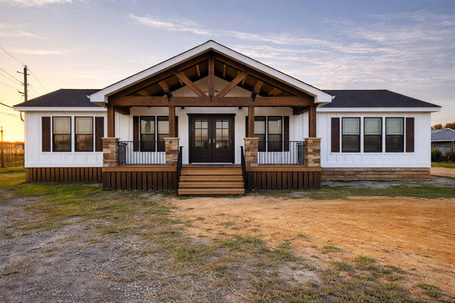 A single-story house with a modern rustic design features a central porch with wooden beams and stone accents. The white exterior and dark shutters create a cozy, welcoming look under a partly cloudy sky.