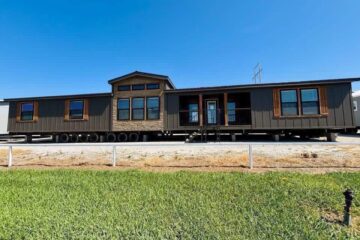 A large manufactured home sits on multiple wheels under a clear blue sky. It features dark siding, several windows, and a centered porch area. The foreground shows a grassy lawn.