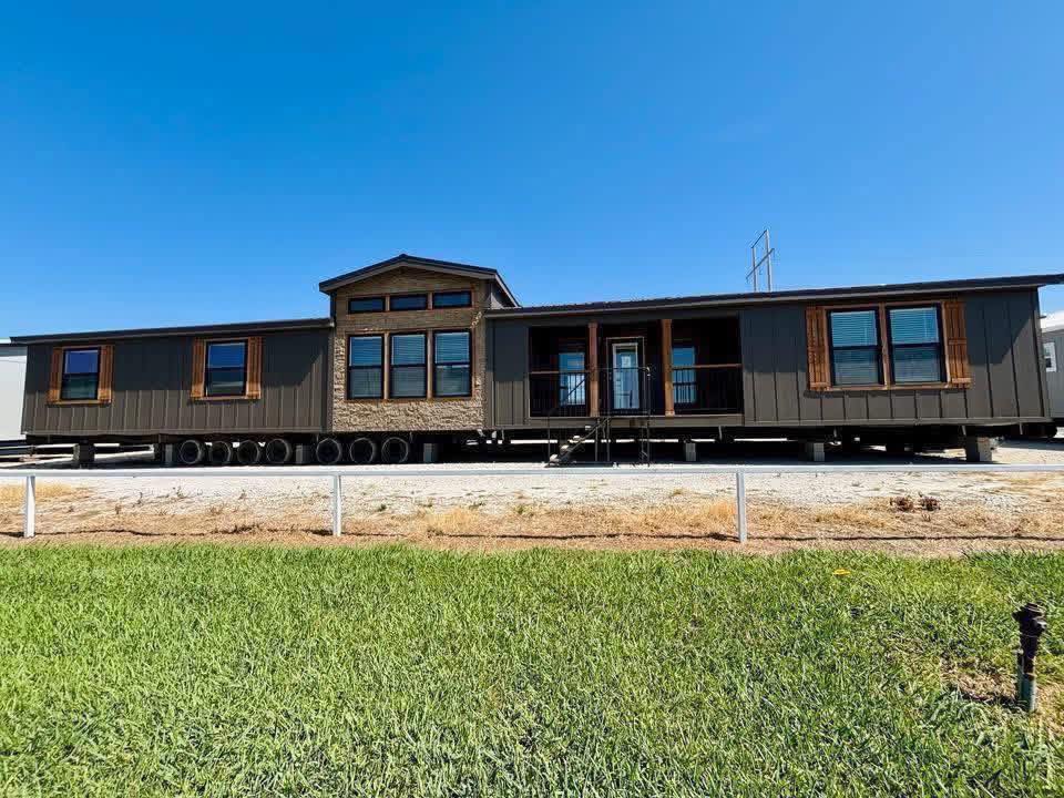 A large manufactured home sits on multiple wheels under a clear blue sky. It features dark siding, several windows, and a centered porch area. The foreground shows a grassy lawn.
