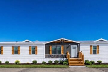 A modern, single-story white manufactured home with wooden shutters and a stone accent facade, set against a clear blue sky. A small porch with steps leads to the entrance, surrounded by neatly arranged shrubs, conveying a tidy, welcoming vibe.