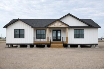 A modern white modular home with black window frames, a covered front porch, and wooden steps, set on concrete blocks against a cloudy sky.