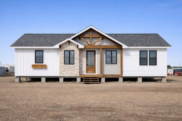 A modern, single-story prefab home with white siding, stone accents, and a wooden door under a peaked roof. It's set in a gravel lot under a clear blue sky.