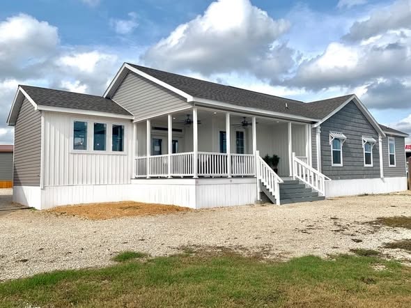 Single-story house with a gray and white exterior, a covered porch, and stairs. It sits on a gravel driveway under a partly cloudy sky, conveying a quaint and peaceful vibe.