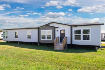 A modern white and gray manufactured home sits on a green lawn under a blue sky with clouds. It features large windows and a small wooden porch.