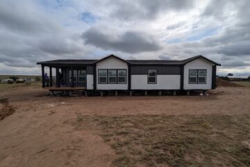 A modern modular home with white walls and dark trim sits elevated on a dirt lot under a cloudy sky, exuding a simple, rustic atmosphere.