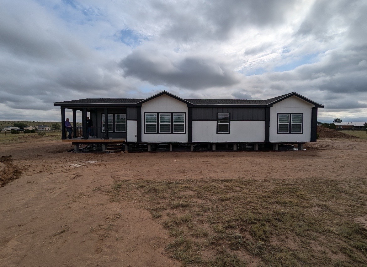 A modern modular home with white walls and dark trim sits elevated on a dirt lot under a cloudy sky, exuding a simple, rustic atmosphere.