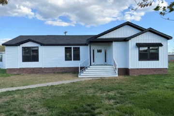 A single-story white house with a black roof and brick foundation, featuring a front porch with stairs and railing, set on a grassy lawn under a partly cloudy sky.