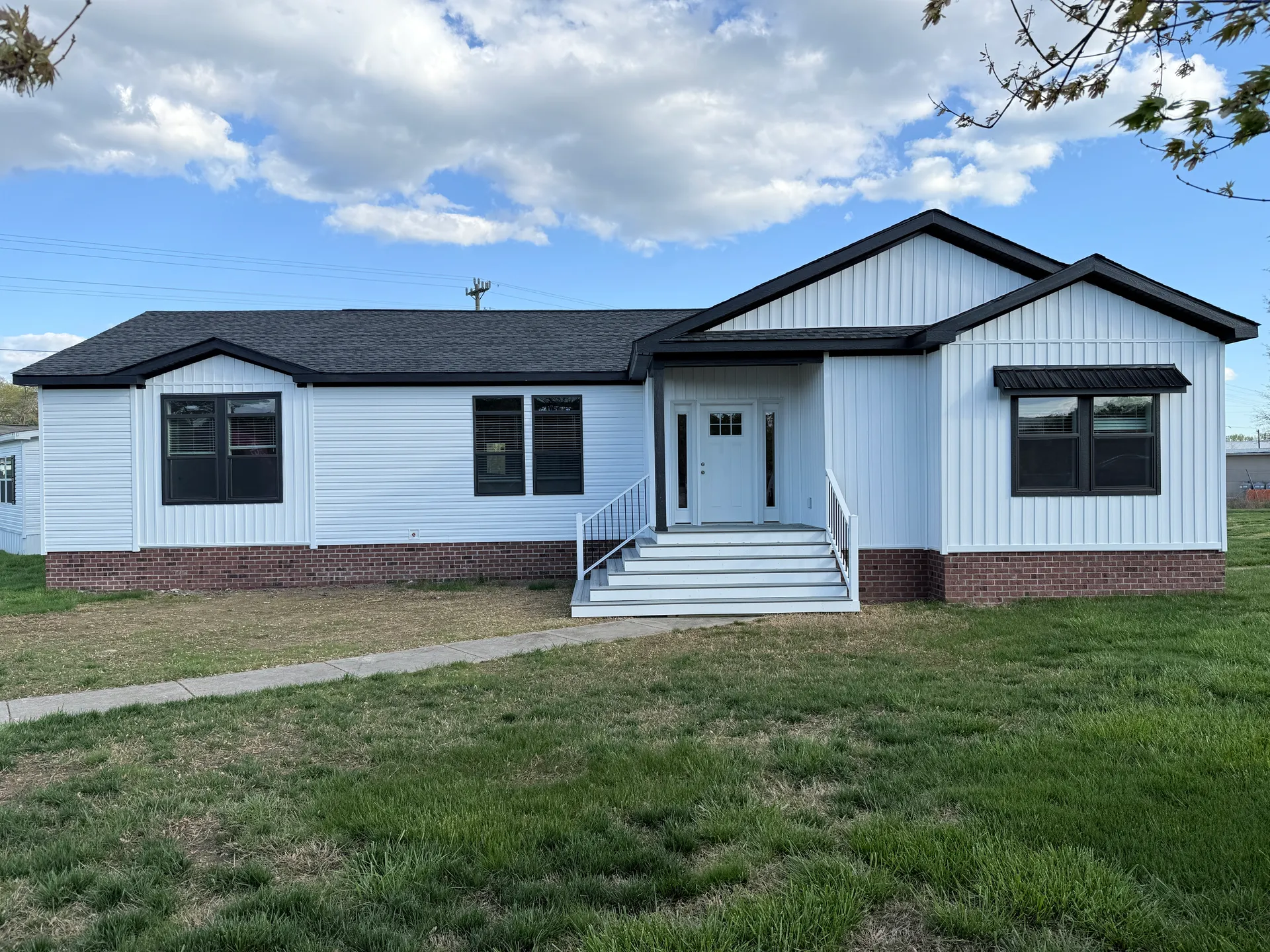 A single-story white house with a black roof and brick foundation, featuring a front porch with stairs and railing, set on a grassy lawn under a partly cloudy sky.